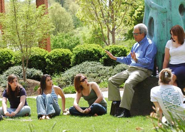 Roger Wilkins rests on the plinth of &quot;Communitas&quot; environmental sculpture on the Fairfax campus of Ƶ. He is speaking to a group of students. It is a beautiful sunny spring day, and students listen attentively as they are seated on the ground. Wilkins is wearing khaki trousers, a blue oxford shirt, and sunglasses.